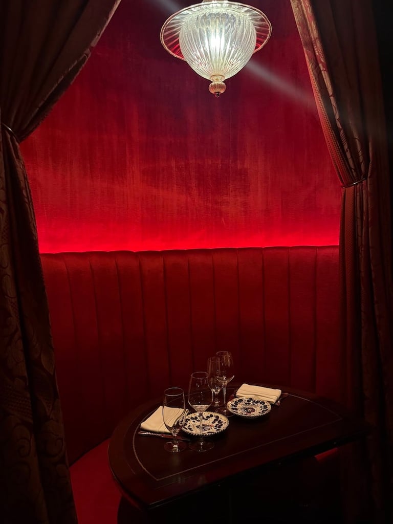 Intimate booth with deep red velvet walls and curtains, featuring a vintage glass ceiling lamp above a table set with appetizers and glassware.