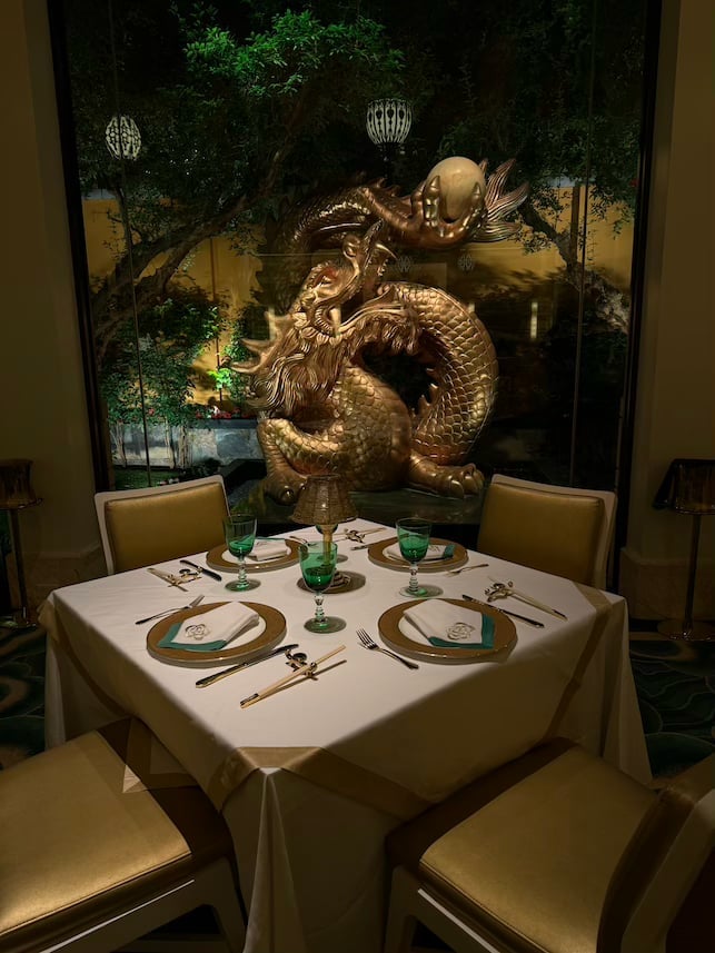 Elegant dining table set for two with green accents, backdrop of large golden dragon sculpture illuminated against dark foliage and decorative lanterns
