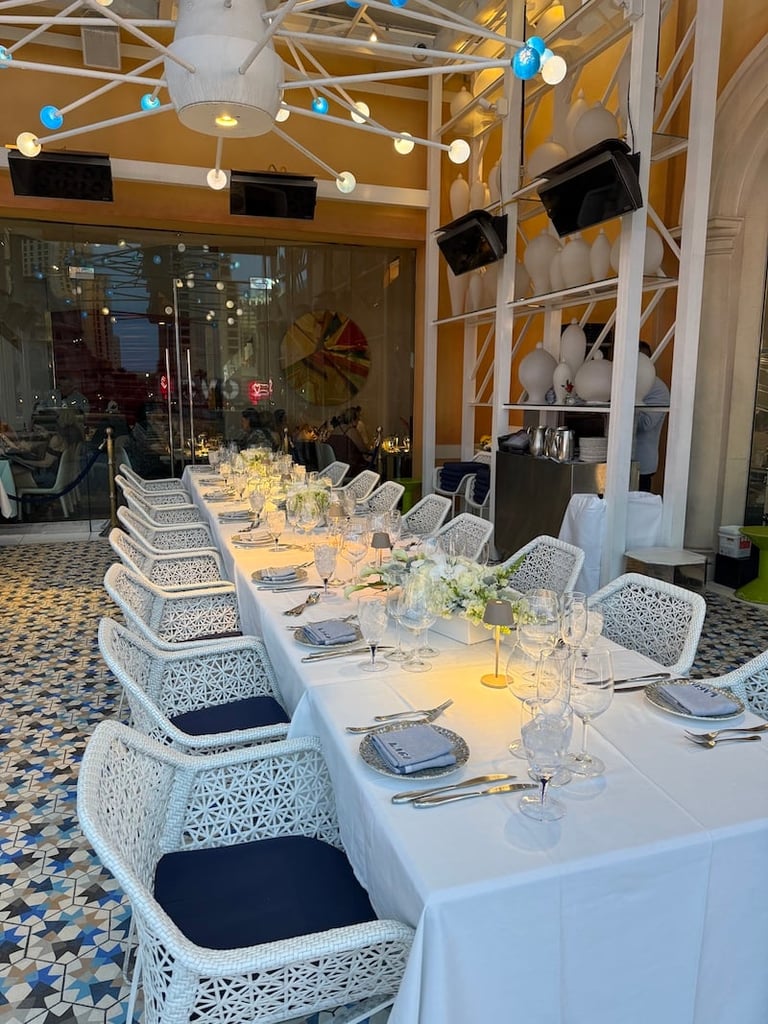 Elegant banquet hall with long white-draped table, white latticed chairs, place settings, floral centerpieces, and blue and white hanging lights above.