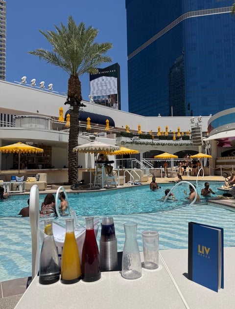 Luxury hotel pool with turquoise water, yellow umbrellas, palm tree, and beverage service in foreground with blue buildings behind