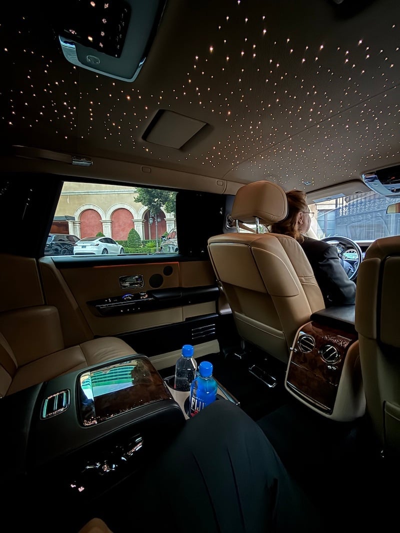 Interior of a luxury vehicle with tan leather seats, starry ceiling lights, rear entertainment screen, and blue water bottles on the console