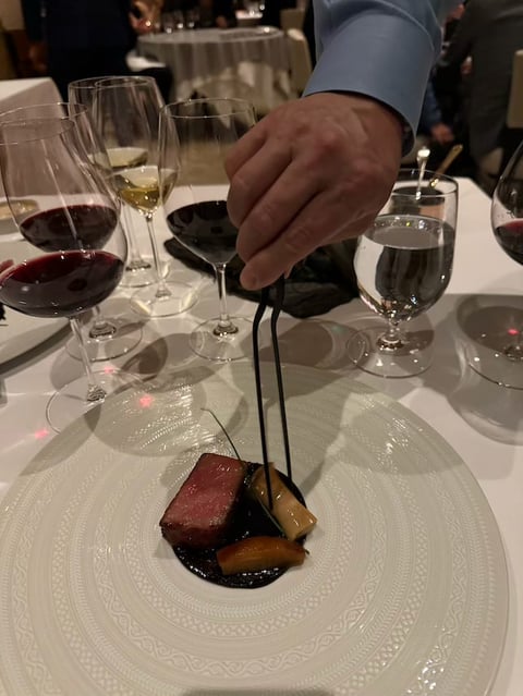 Person in blue sleeve presenting plated dessert with dark sauce and garnish at fine dining table with wine glasses