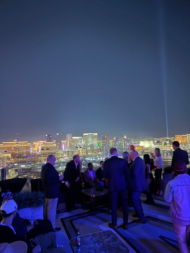 Group of people on an observation deck at night overlooking a brightly lit cityscape with tall buildings and a light beam in the sky