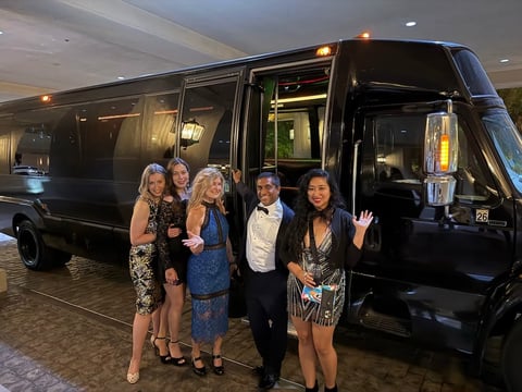 Five people in formal attire standing in front of a black limousine in an indoor garage setting