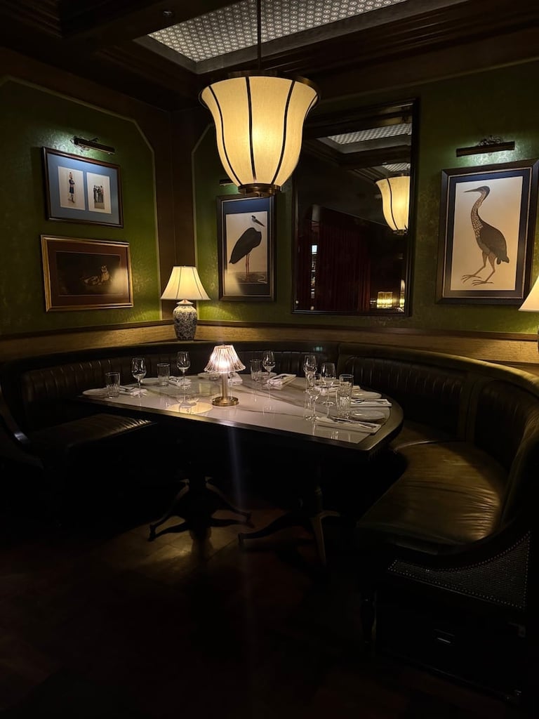 Elegant dining room with green walls, vintage art prints, hanging pendant light, black booth seating, and formally set round table with place settings.