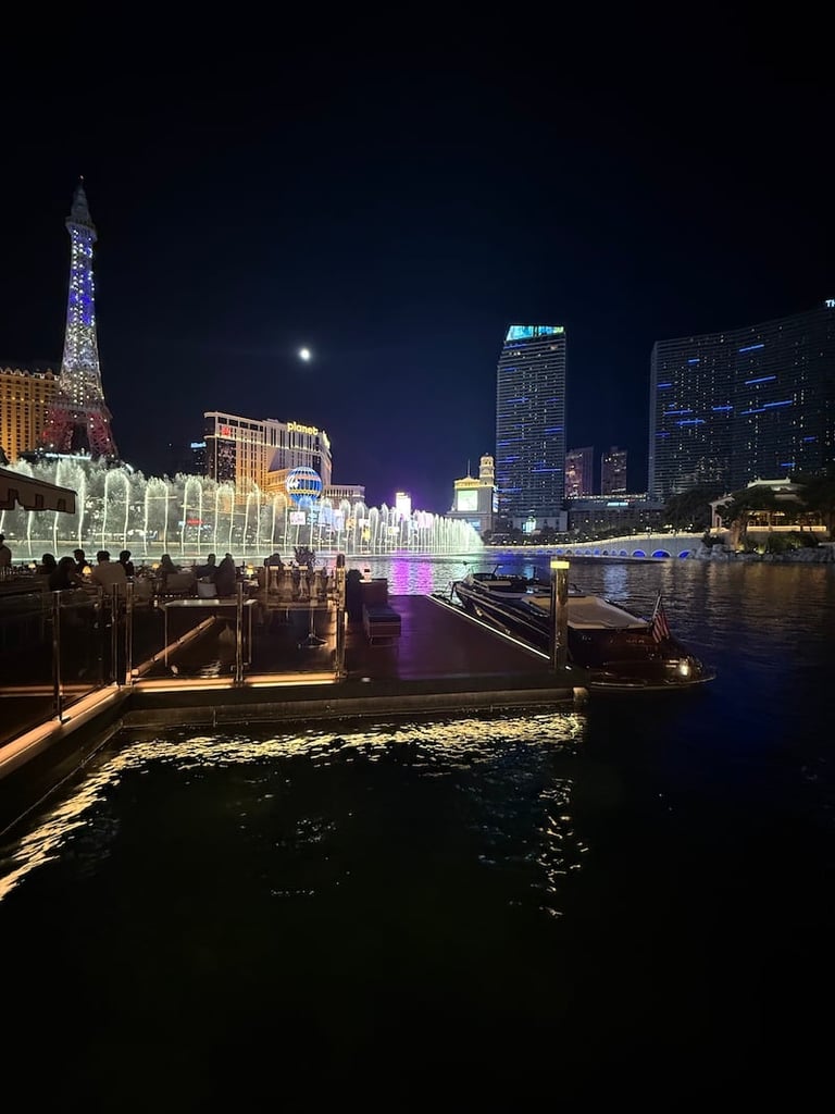 Night cityscape of Las Vegas Strip showing illuminated buildings, Eiffel Tower replica, and water canal with colorful reflections.
