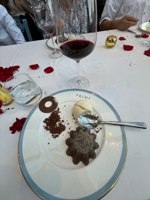 Plated chocolate dessert with chocolate crumbles and star-shaped pastry, accompanied by red wine glass and rose petals on white tablecloth.