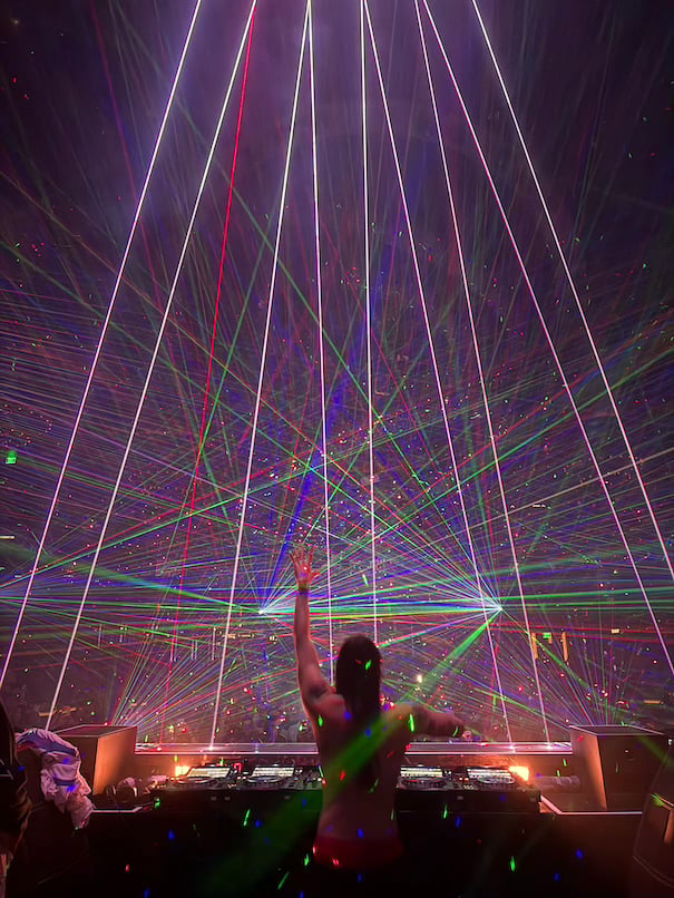 DJ performing under a vibrant laser light show with purple, pink, and green beams radiating across a dark venue with silhouetted crowd