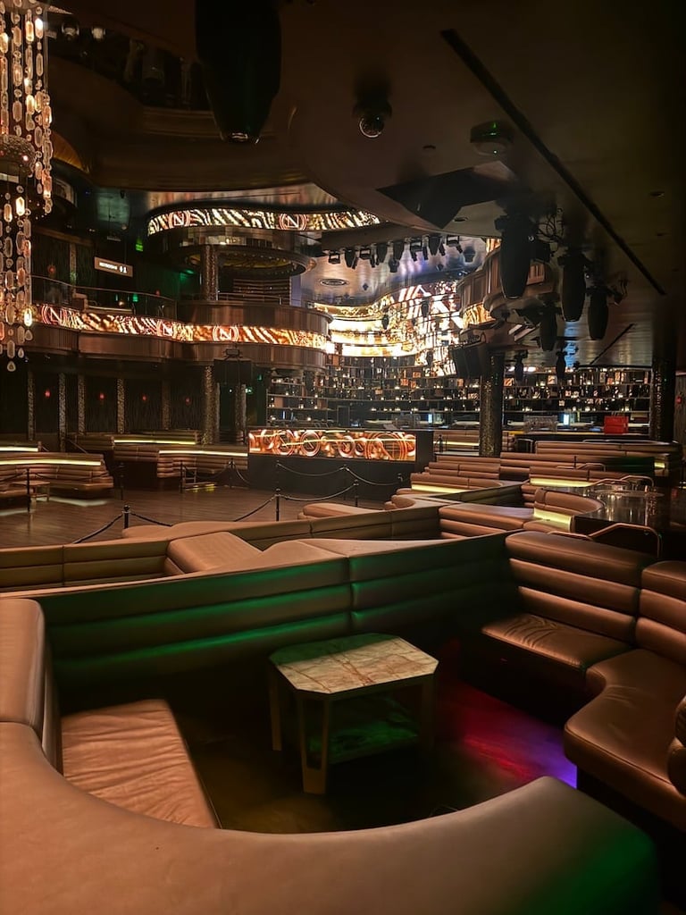 Interior of an upscale nightclub with curved green and brown leather booths, an illuminated stage with neon signage, and dramatic lighting