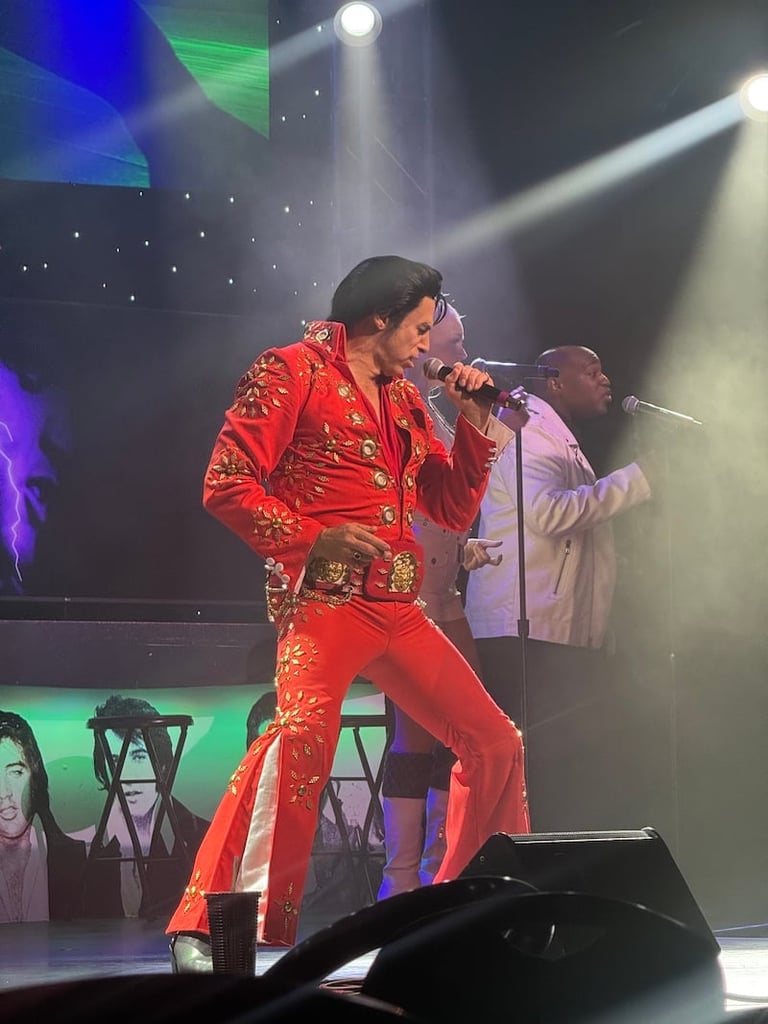 Two performers on stage singing into microphones, one wearing a red sequined jumpsuit and the other in a light-colored jacket, with dramatic stage lighting