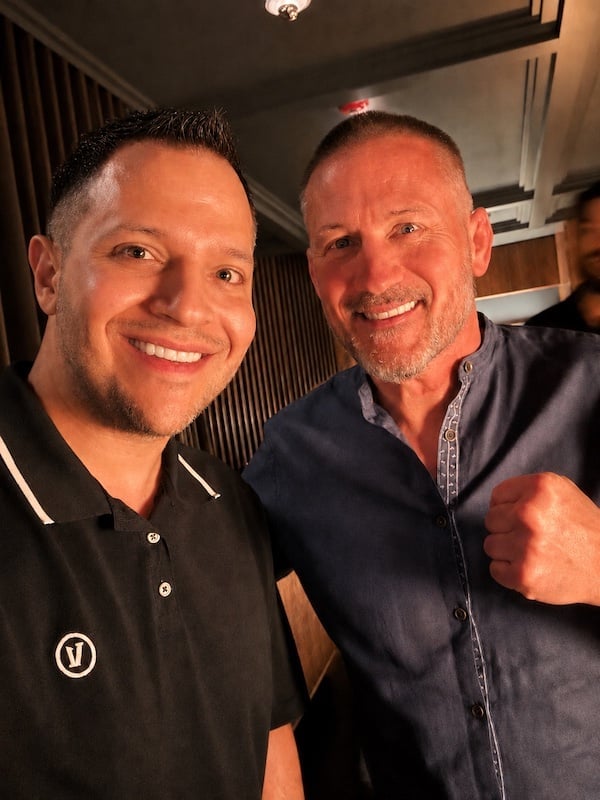 Two men smiling at the camera in an indoor setting, one wearing a black polo shirt and the other a dark blue denim shirt.