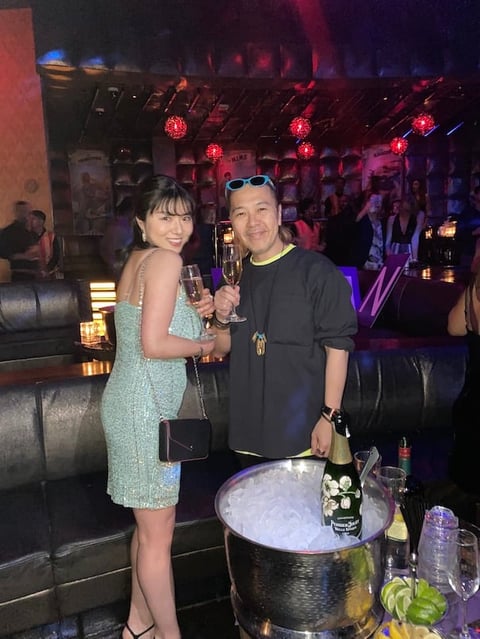 Two people posing at a nightclub holding drinks, with neon red lights and ice bucket visible in the background