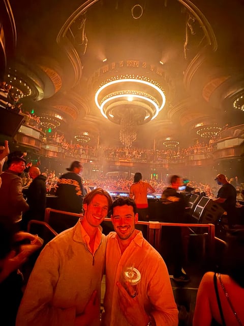 Two men smiling at camera inside ornate concert venue with golden lighting and large illuminated chandelier overhead