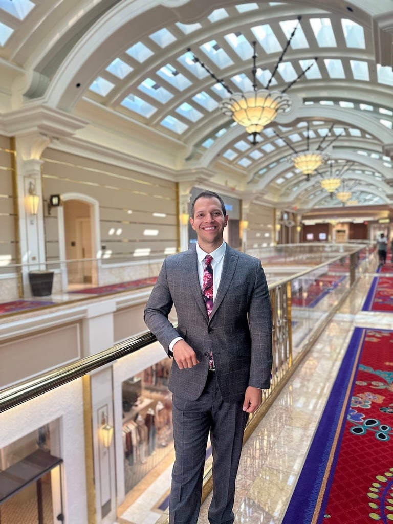 Man in gray suit standing on a balcony inside an elegant interior with arched glass ceiling and decorative lighting