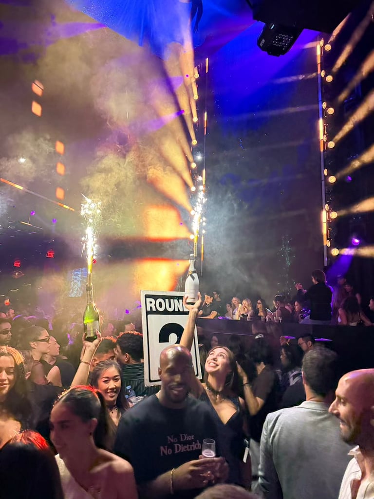 Crowded nightclub with dancing audience, stage pyrotechnics, blue and yellow lighting, and raised bottle in center of crowd.