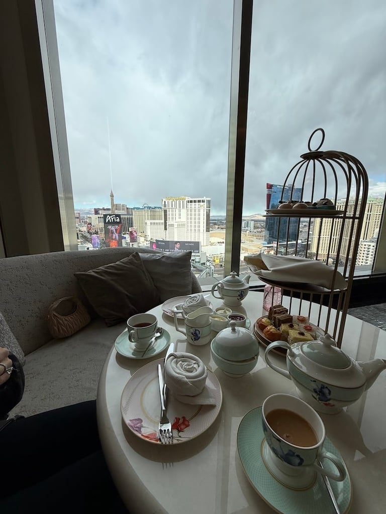High-rise window view overlooking a city skyline with a tea service set on a table, including teacups, teapot, pastries, and a decorative birdcage.