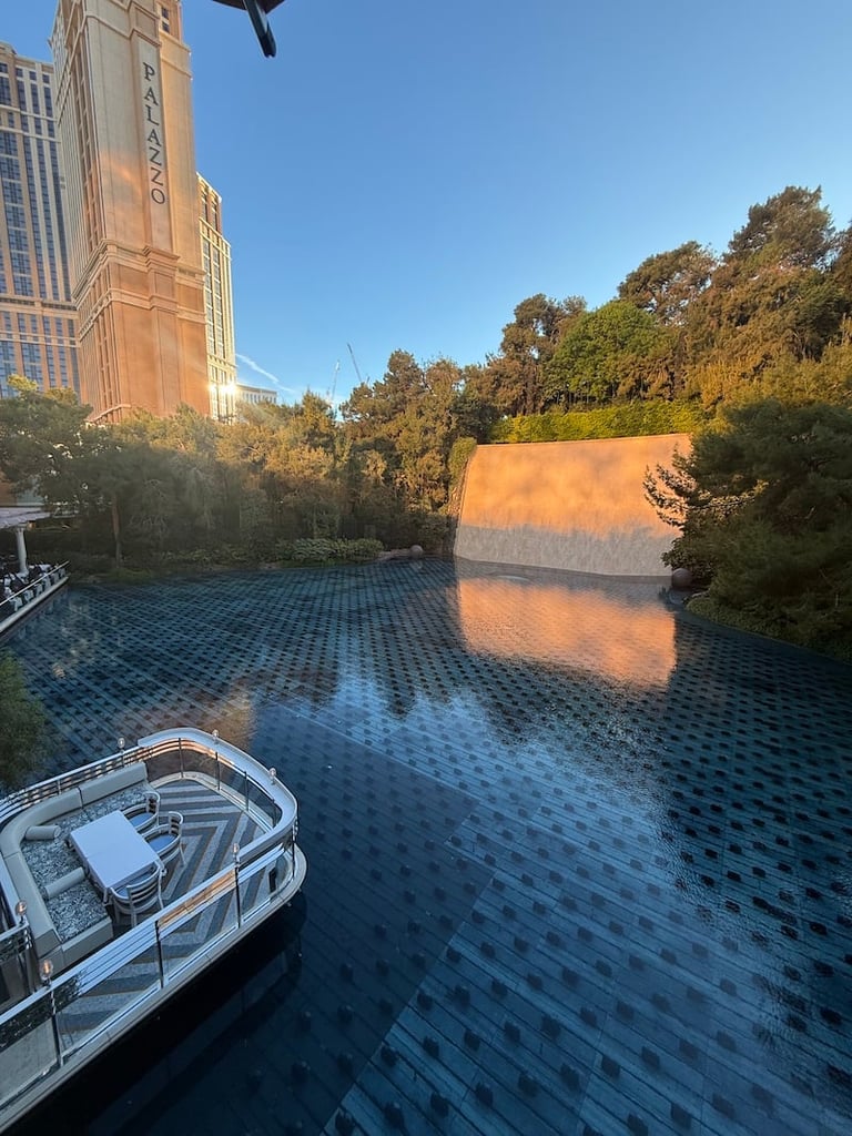 Urban waterway with moored boat, reflecting large orange building, trees, and city skyline at sunset.