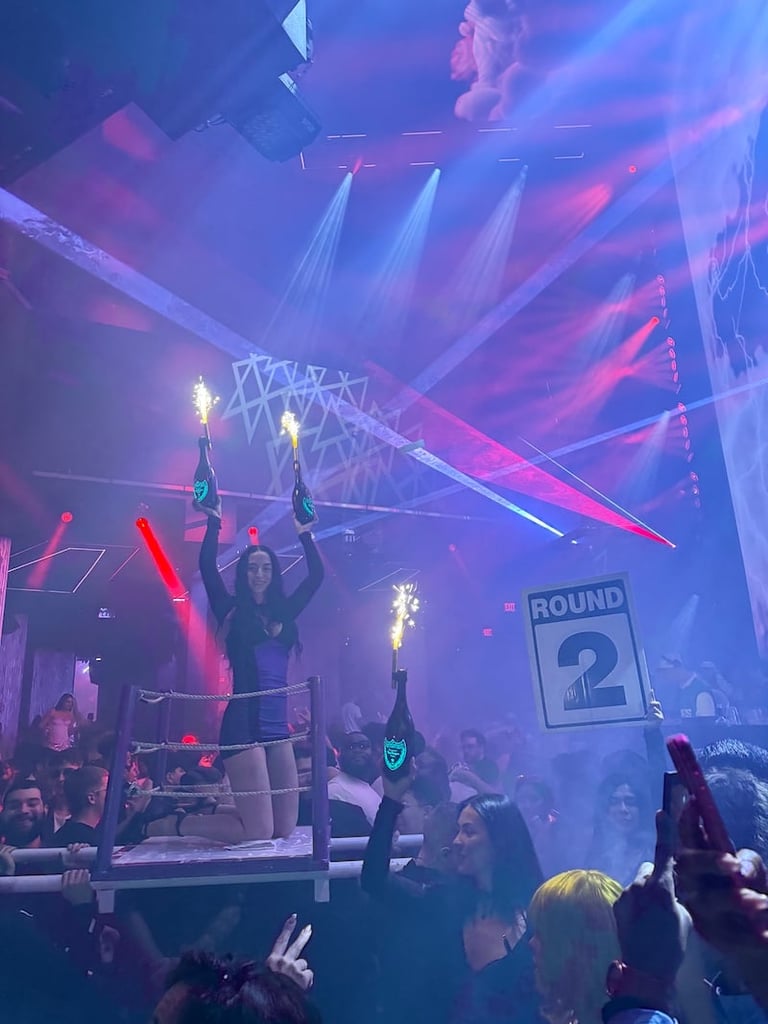 Nightclub stage with performers on elevated platform, blue and pink laser lights, round 2 sign, and crowd dancing below