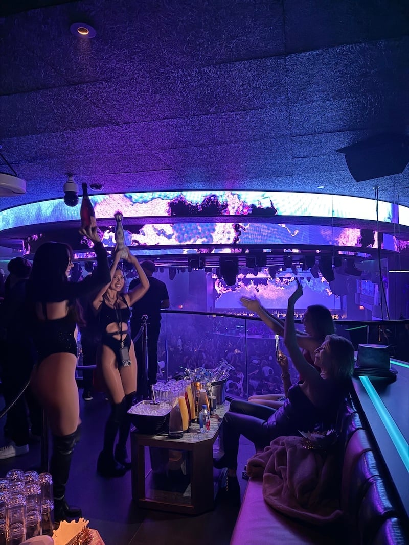 People dancing and socializing in a nightclub with purple and blue neon lighting and a large curved display screen above the bar.
