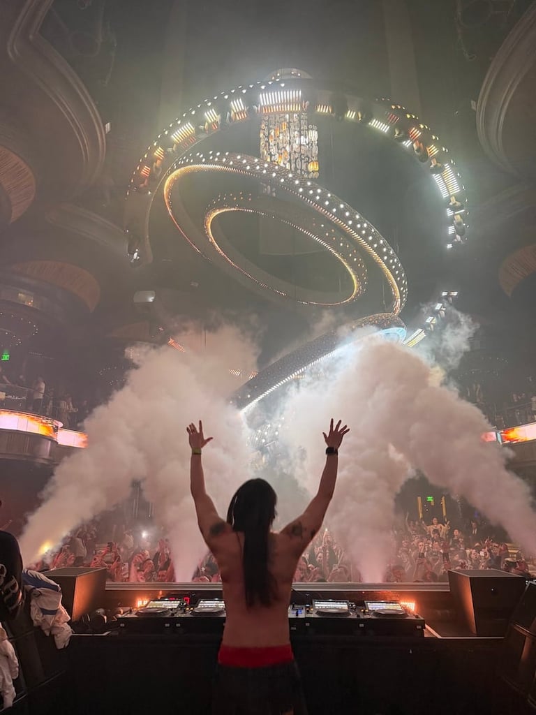 Person with raised arms facing massive illuminated stage with spiraling lights and white pyrotechnic fog effects at concert venue.