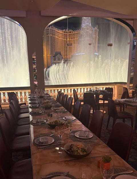 Upscale dining room with long table set for dinner, overlooking illuminated Bellagio fountains through large windows