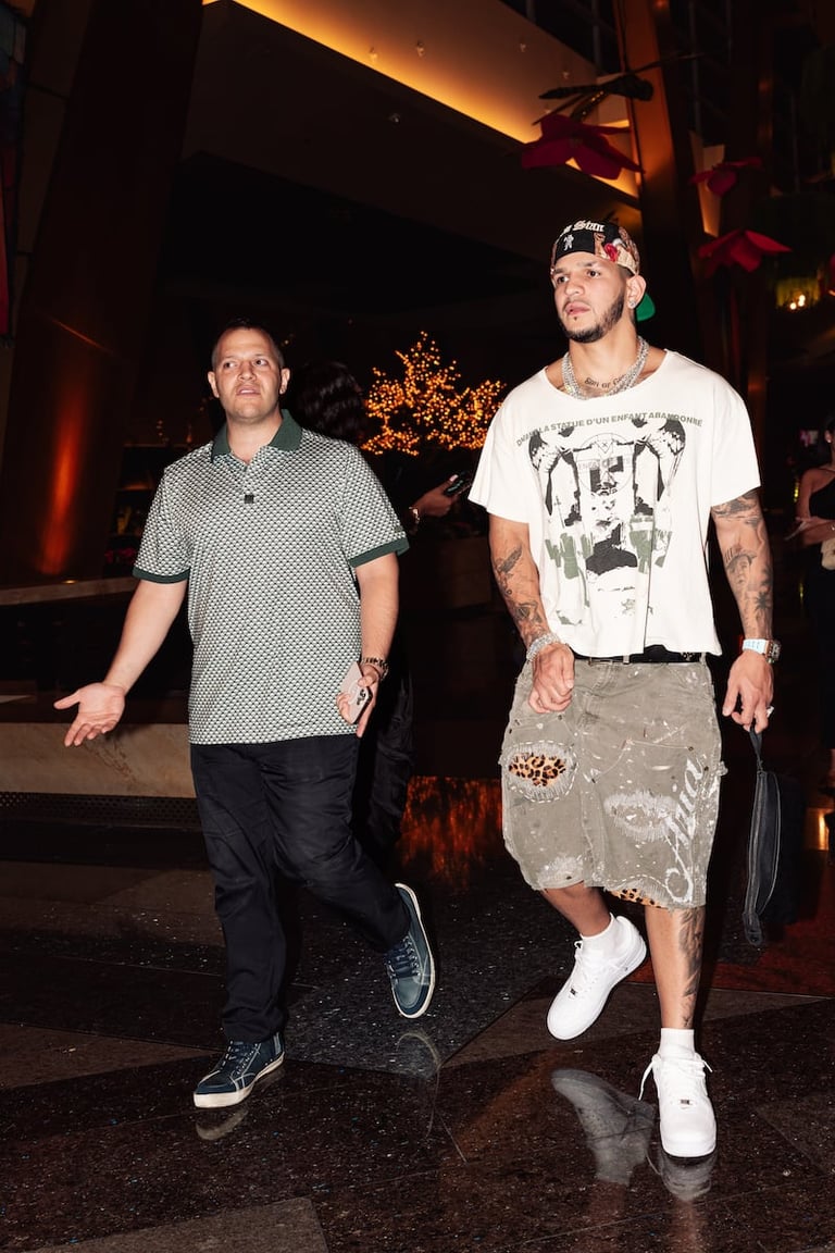 Two men walking together at night outside a venue with warm lighting, one wearing a patterned shirt and the other in a white graphic tee.